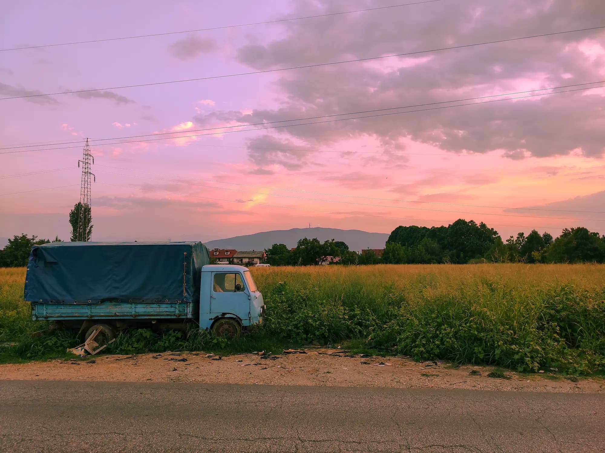 An abandoned turquoise flatbed truck with a canvas canopy, parked in an overgrown field beside a road under a pink and orange sunset.