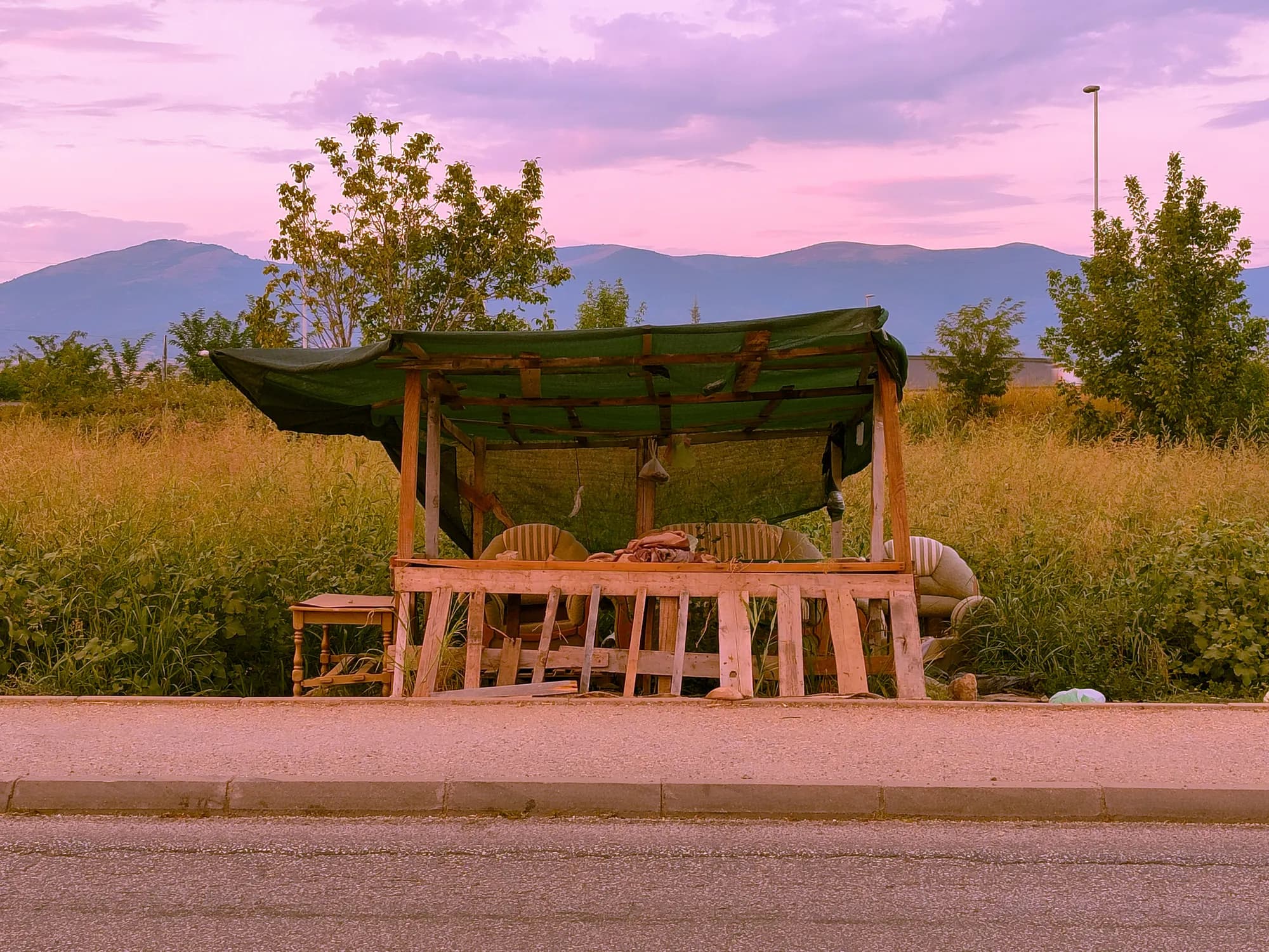 An empty roadside stall built from raw timber under a green tarpaulin, standing in tall grass with mountains behind it at dusk.