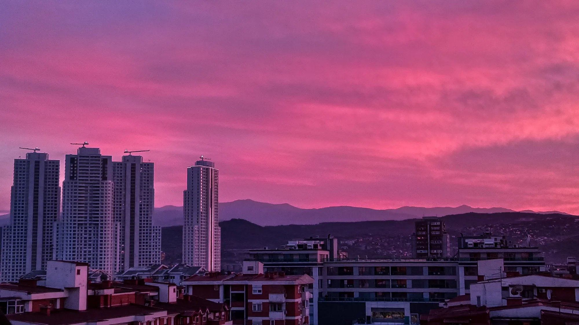 A city skyline at dusk under magenta cloud, with unfinished high-rise towers on the left and a mountain ridge behind.