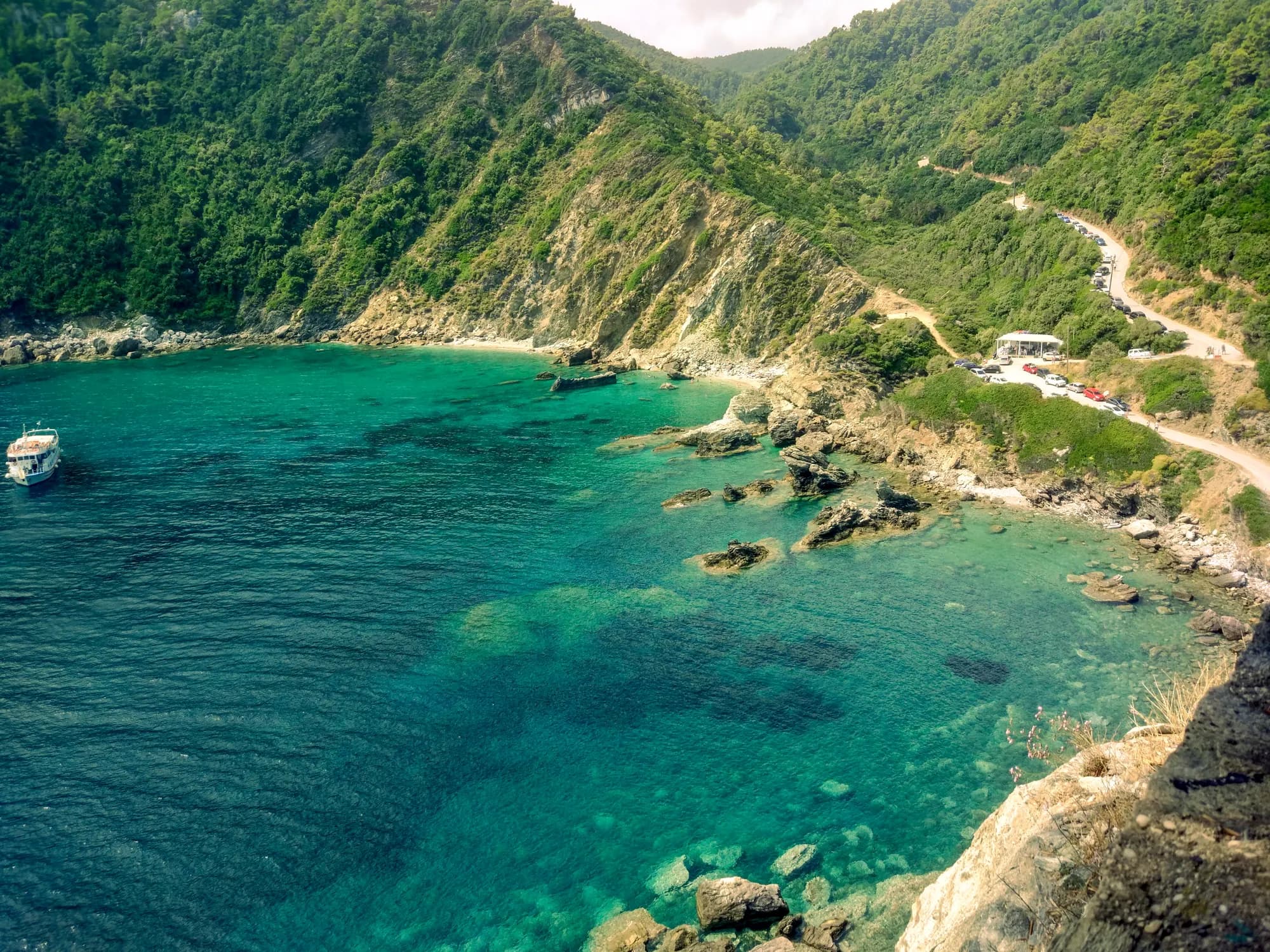 A cove seen from above: turquoise water over pale rock, wooded cliffs, a small boat, and a road switchbacking down to the shore.