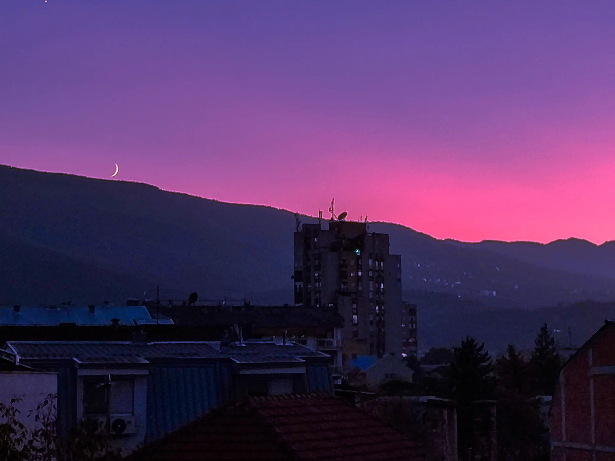 A crescent moon over a dark mountain ridge, under a magenta band of sky, with a tower block and tiled rooftops in the foreground.