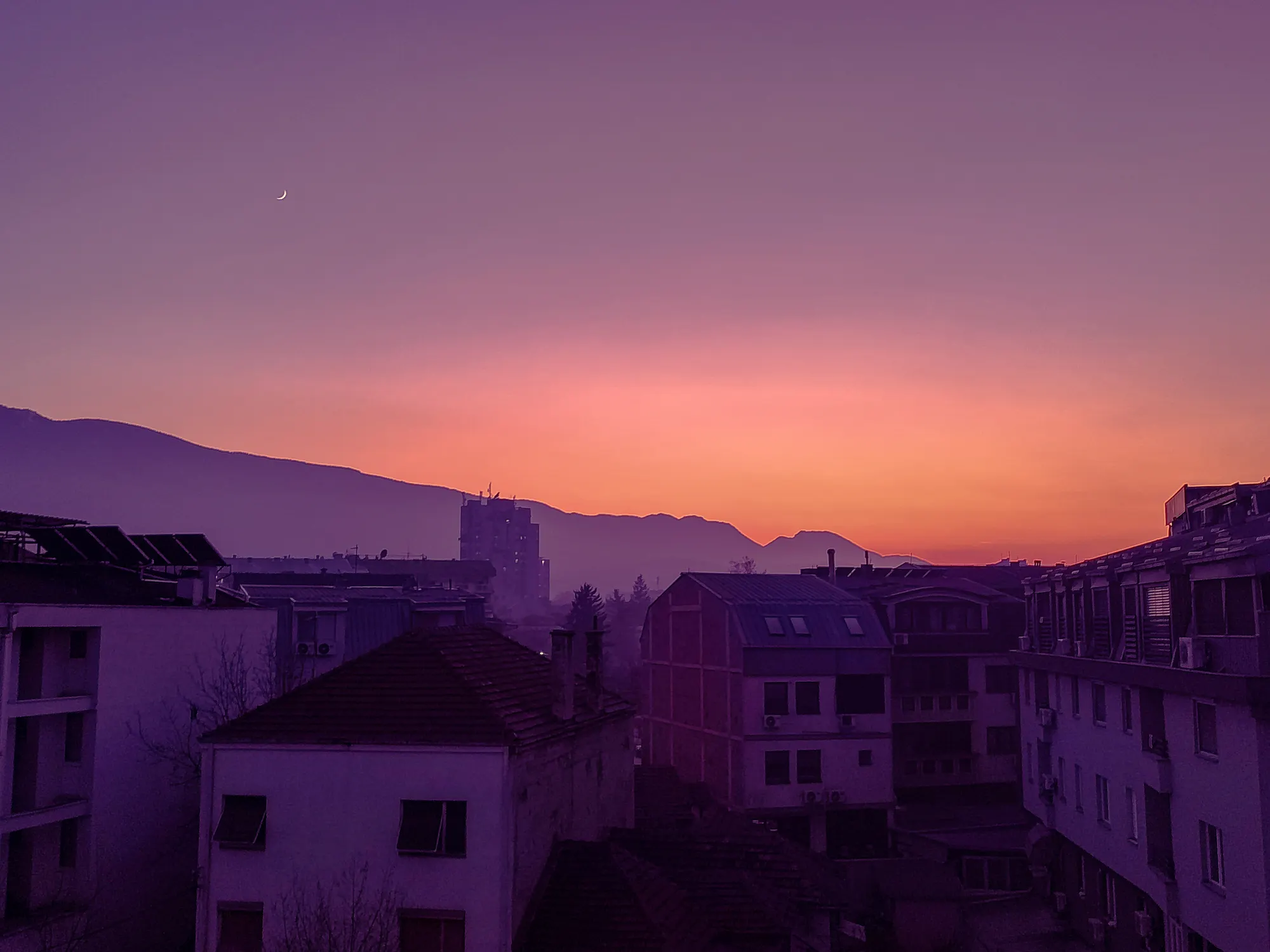 Rooftops of a hillside town in violet haze, with an orange strip of afterglow along the horizon behind them.
