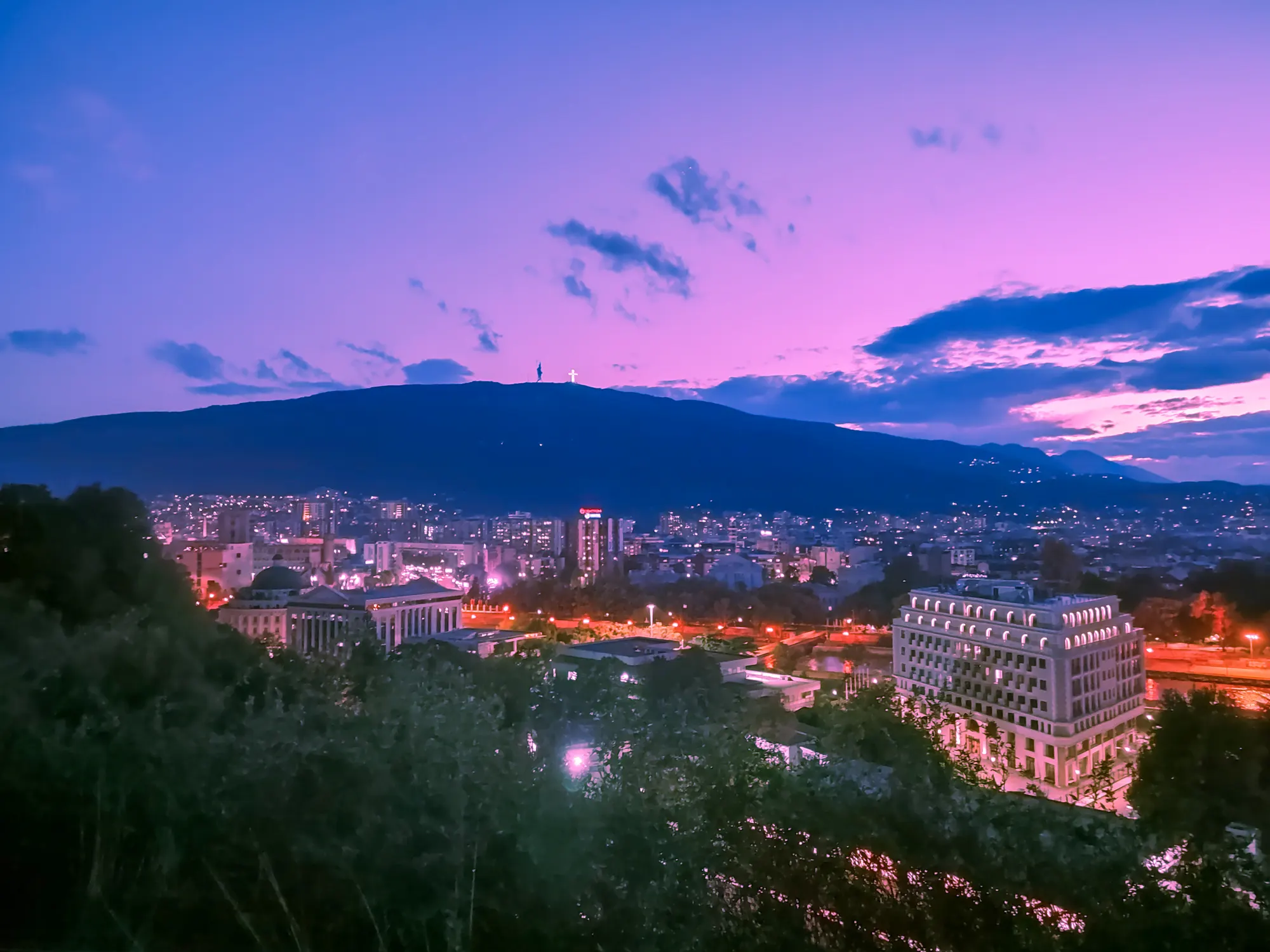 Skopje at dusk from a hillside: lit streets and buildings below, Mount Vodno on the horizon, tree canopy across the bottom of the frame.