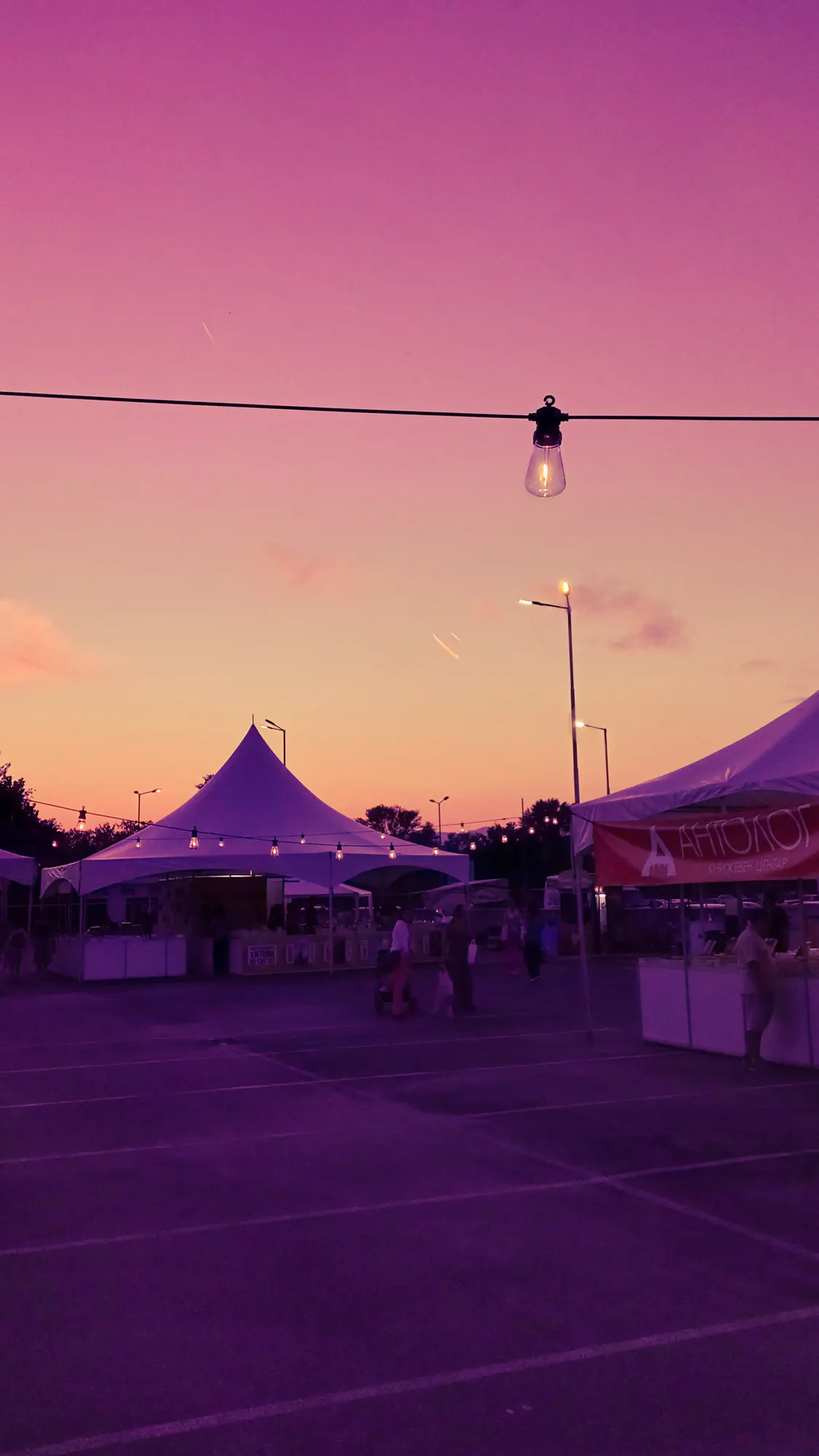 A night market at sunset: white peaked tents, people between the stalls, and a single lit bulb hanging from a wire across the top of the frame.