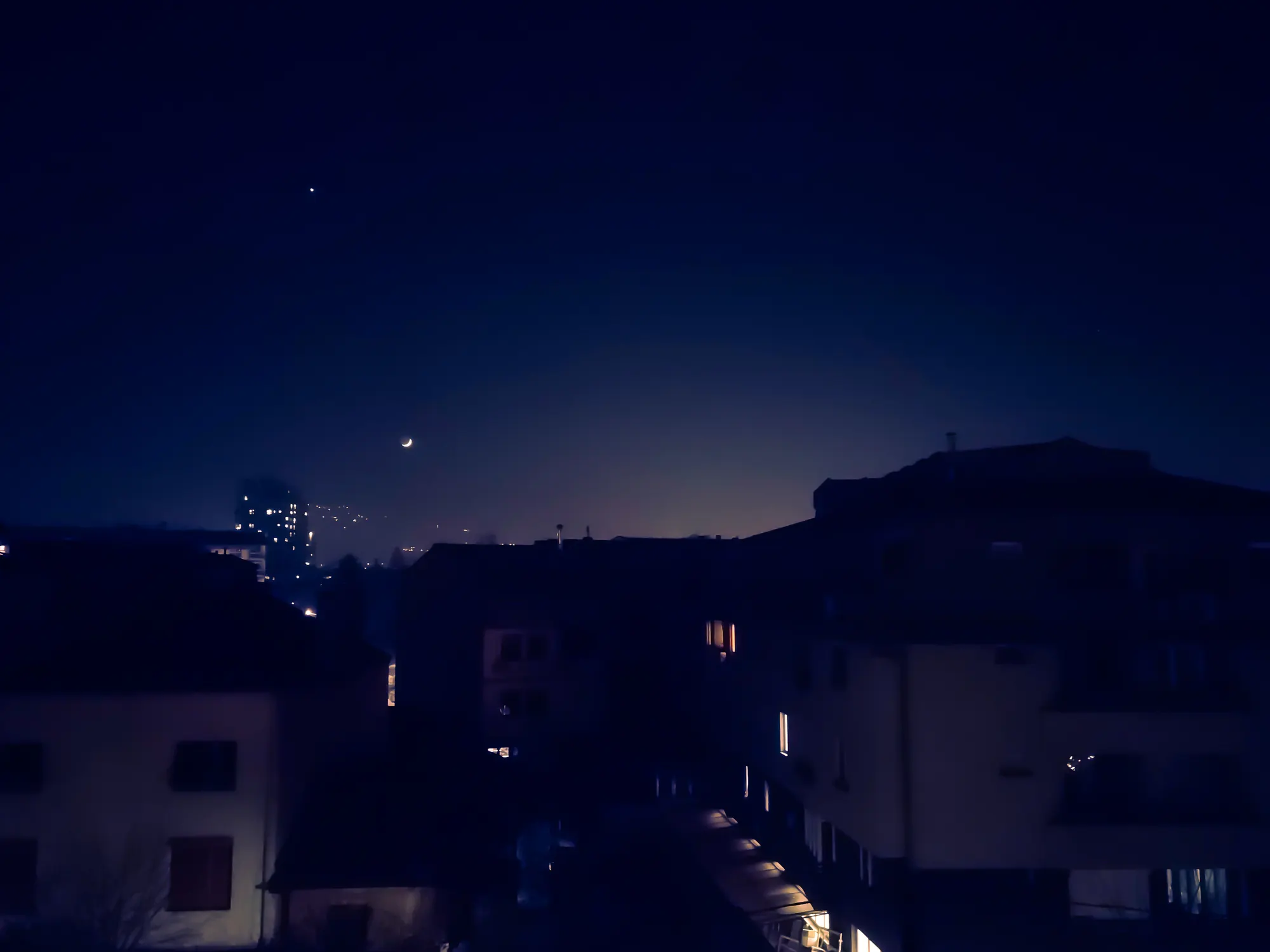Rooftops in near-darkness under a deep blue sky, a faint crescent moon low over the hills and a few lit windows.