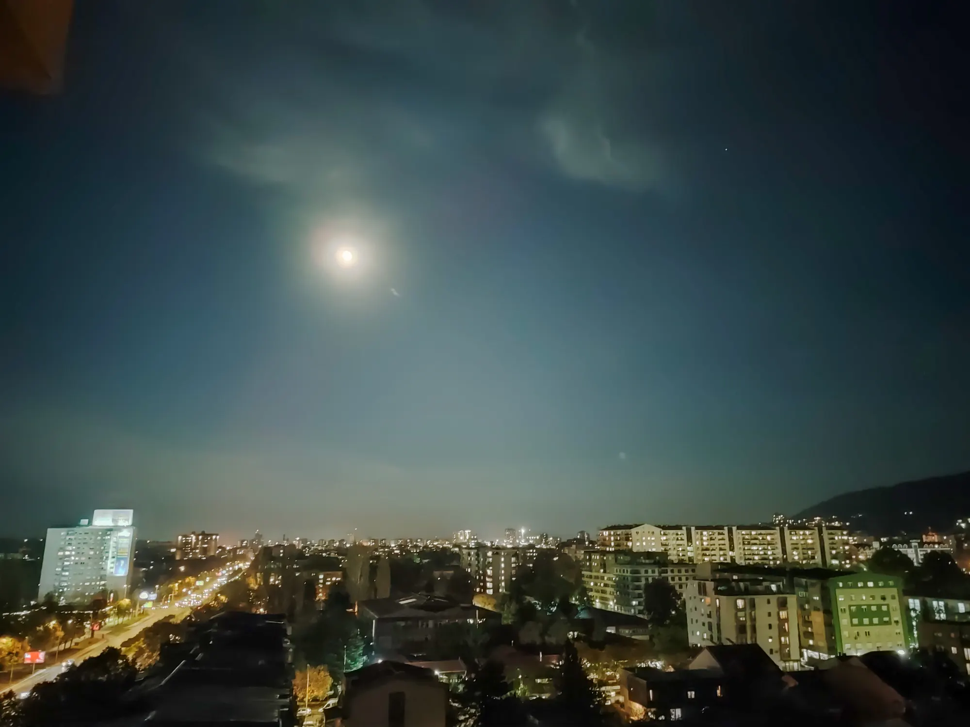 A bright moon high over a city at night, with a lit avenue running between blocks of flats toward distant hills.