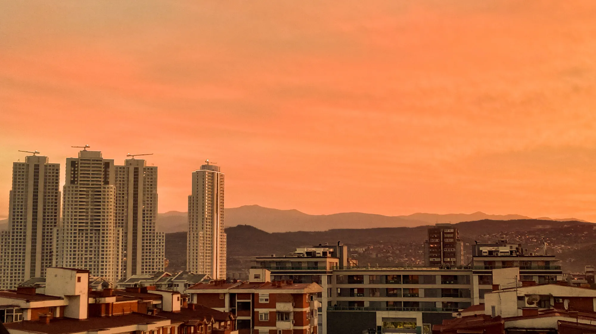 An orange sky over low rooftops, with a pair of pale high-rise towers standing against the mountains.