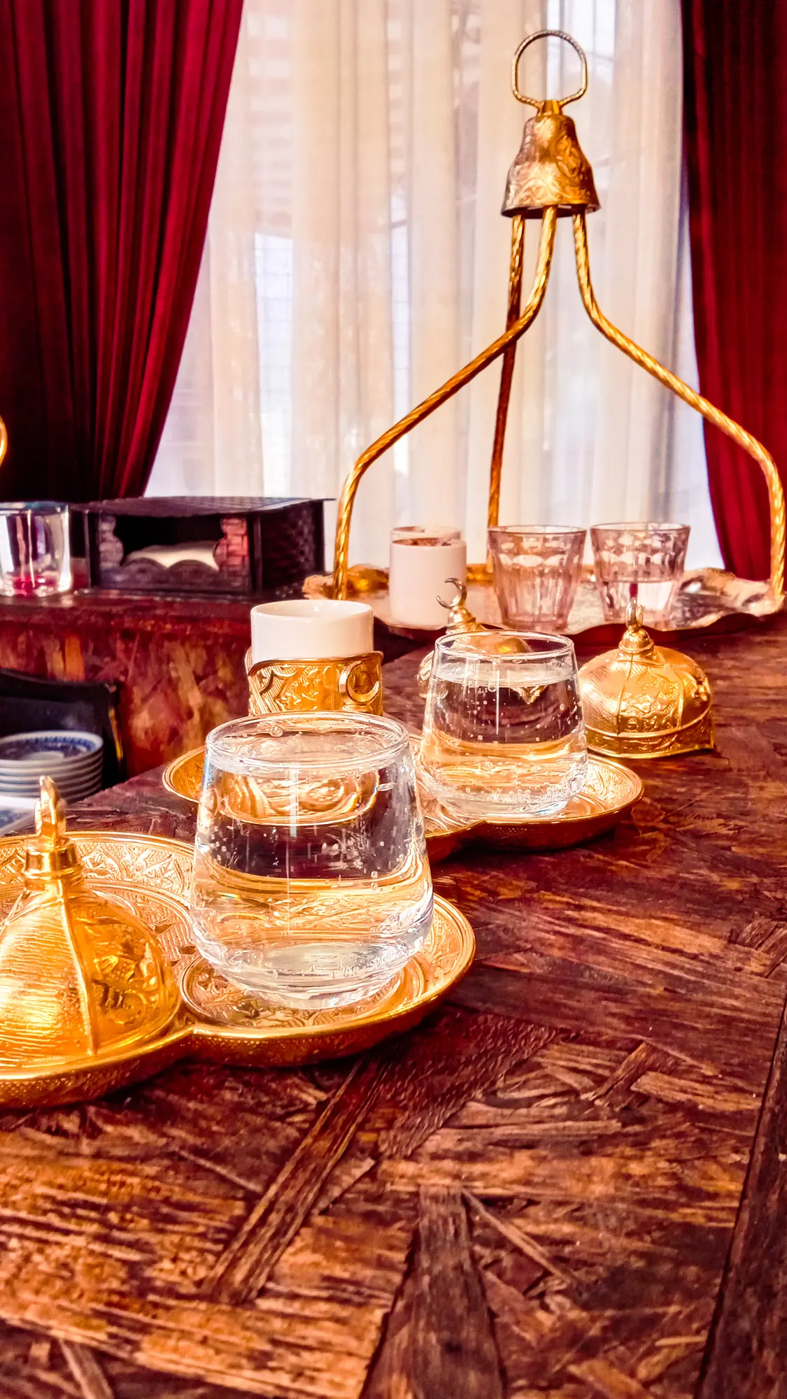 A gilded tray of tulip-shaped tea glasses and lidded sugar bowls on a wooden table, lit warm against red curtains and white sheers.