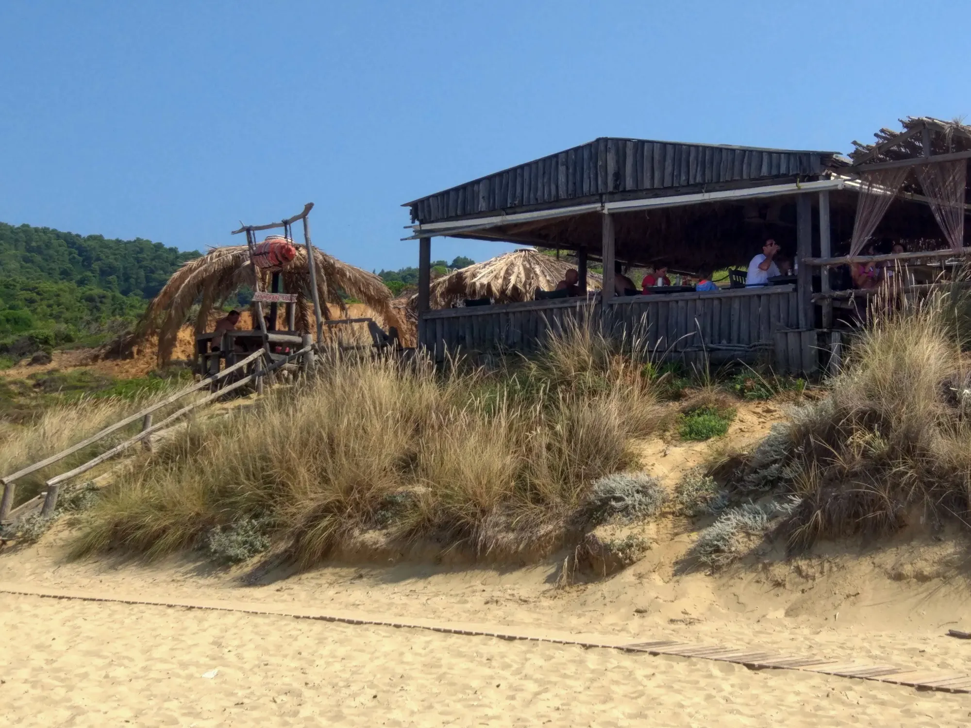 A beach in full daylight: dune grass on the sand, a timber beach bar on the rise above it, thatched parasols and a clear blue sky.