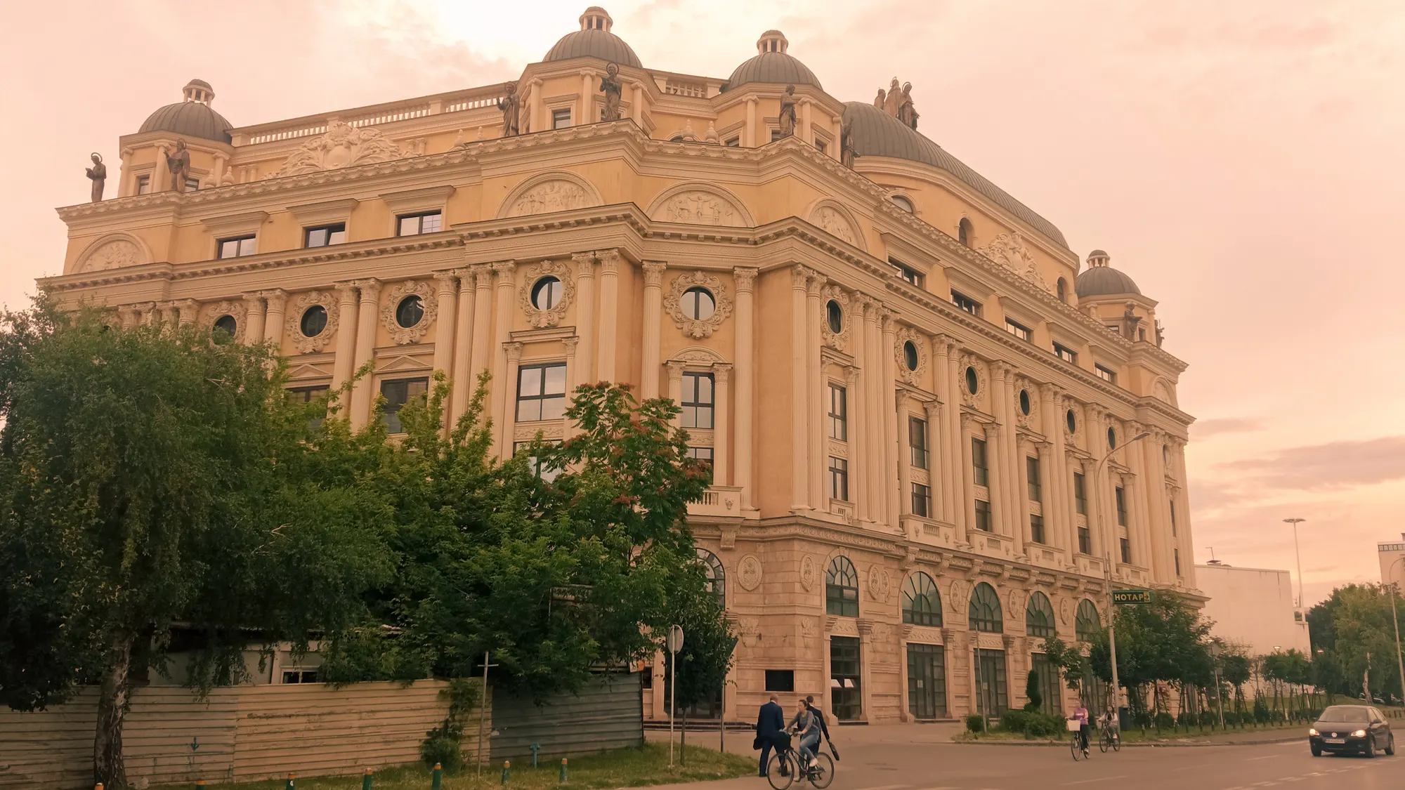 A grand neoclassical building with domed corners and arched windows, photographed from across the street at sunset as two cyclists pass.