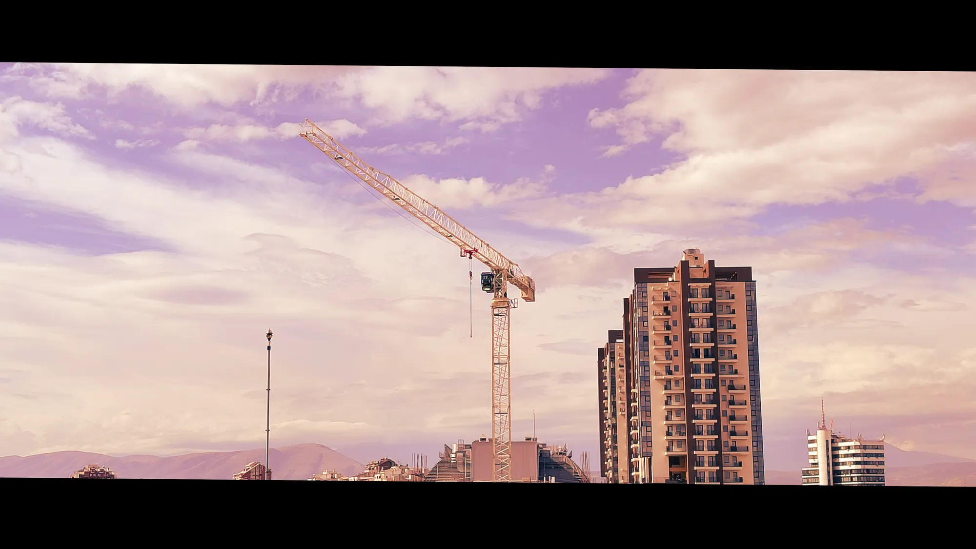 A tower crane beside an unfinished high-rise, both dark against lavender and pink cloud.